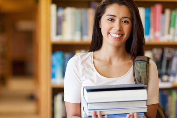 Universitaria en una biblioteca sonriendo motivada y segura de que podrá cursar sus estudios superiores gracias a la Fiducia - Universitas.